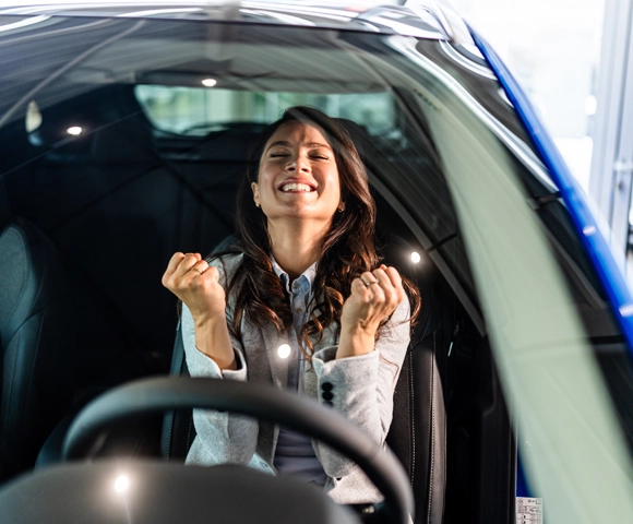 Excited woman making a fist gesture, celebrating a new car inside an automobile dealership. Concept of achievement and success